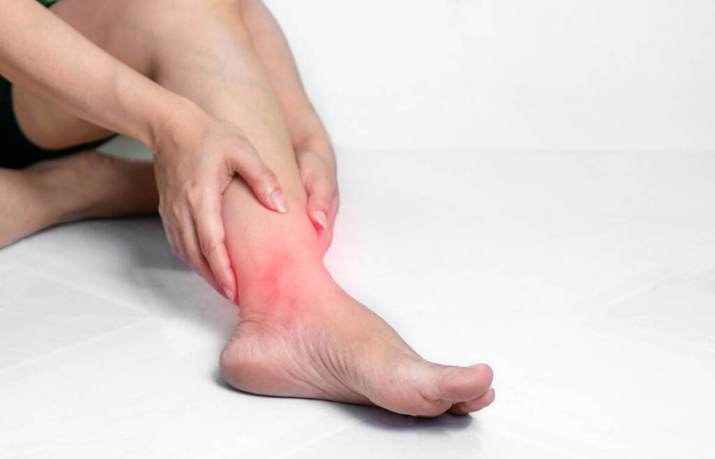 A person sitting on a white surface holding their right ankle, highlighting an area of pain on the side of their foot, depicted by a red overlay.