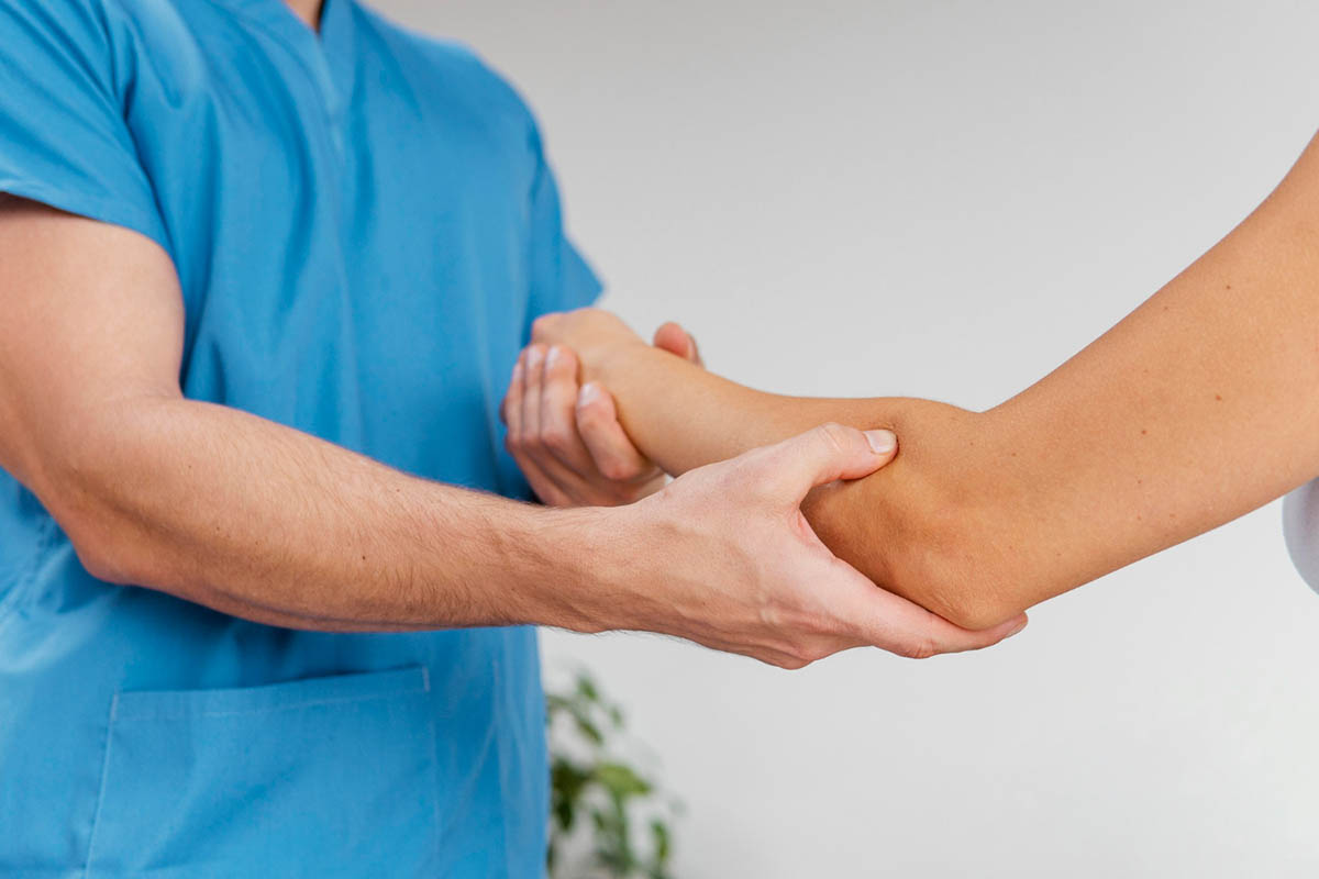 A healthcare professional in blue scrubs examines a patient's elbow, holding it gently with both hands against a neutral background.